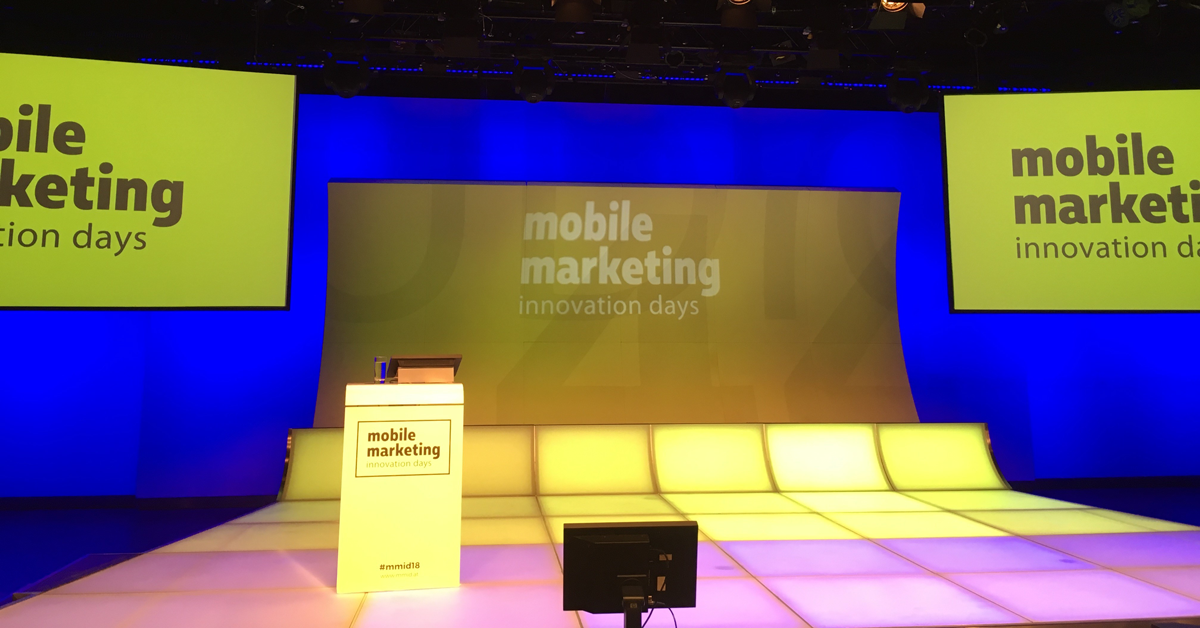 Zu sehen ist die Bühne der Mobile Marketing Innovation Days. Der Boden besteht aus leuchtenden Fliesen, welche im Vordergrund violett, im Hintergrund gelb leuchten. Links auf der Bühne steht ein Rednerpult mit eingebautem Bildschirm. Im Hintergrund ist eine gelbe Plakatwand zu sehen, auf der der Name der Veranstaltung steht.