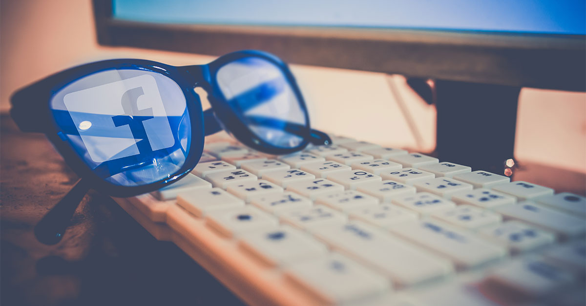Auf einer Tastatur liegt eine Brille, in deren Gläsern sich das Facebook-Logo spiegelt.