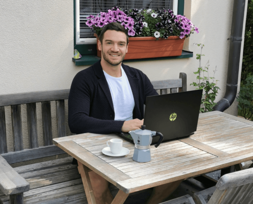 Ein dunkelhaariger junger Mann arbeitet am Laptop und lächelt in die Kamera.